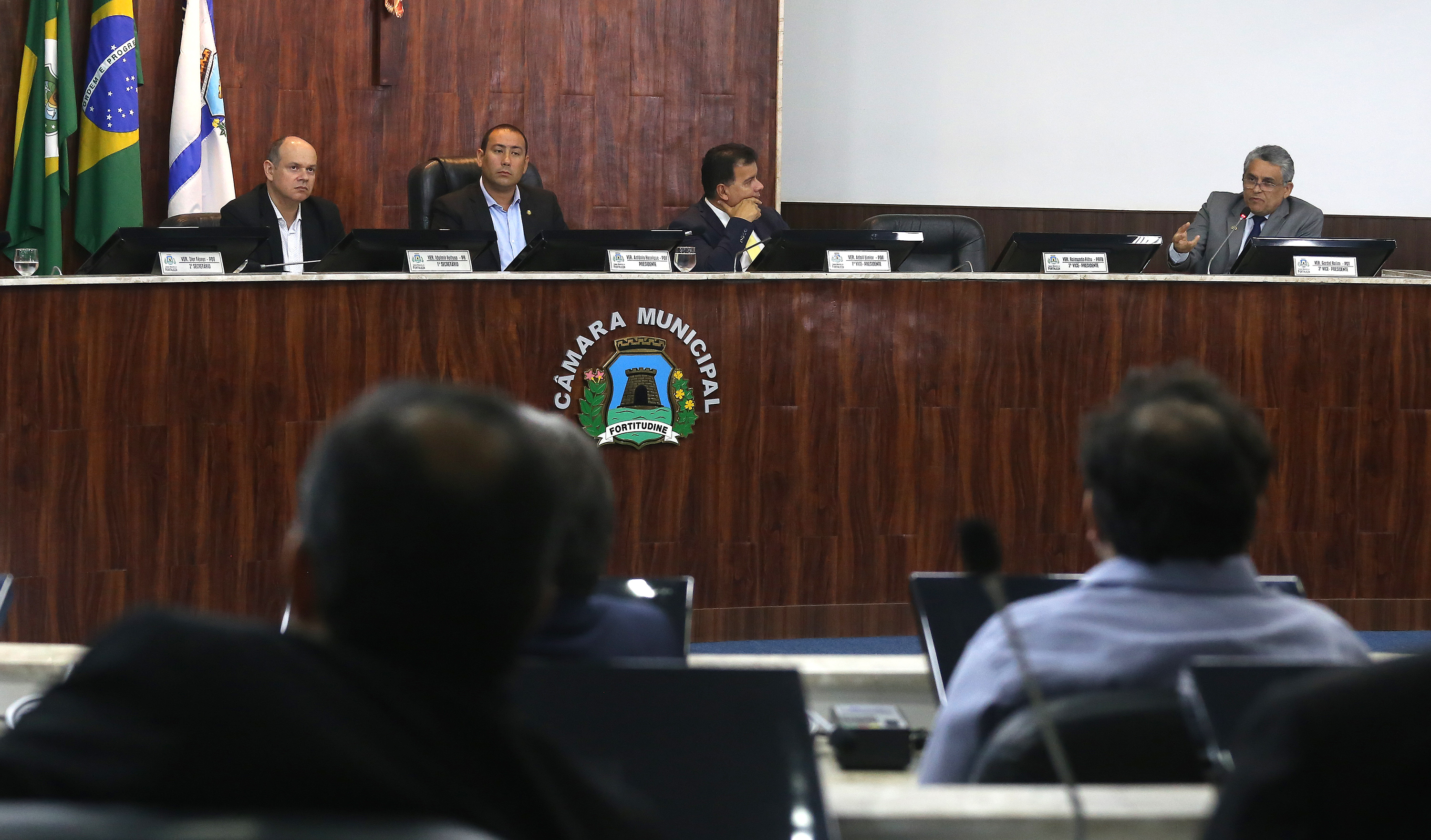 secretário falando ao microfone em mesa com pessoas sentadas ao lado ouvindo